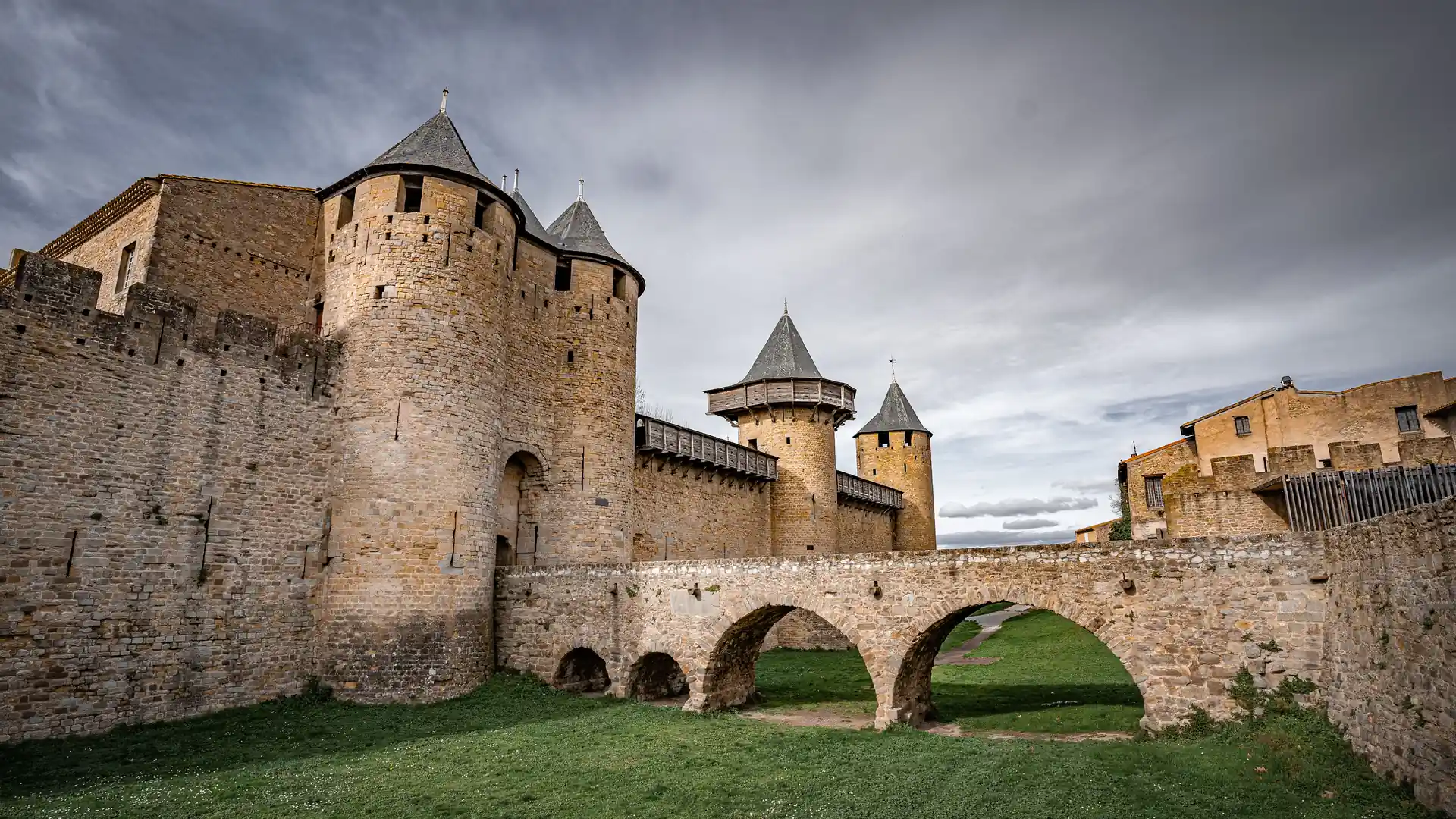 Kulaté věže a kuželové břidlicové střechy hraběcího hradu Château Comtal v Cité de Carcassonne, k němuž vede klenutý kamenný most přes travnatý příkop
